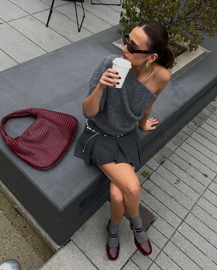 A woman sits on a dark gray outdoor bench, wearing a gray off-the-shoulder knit top, a pleated mini skirt, gray socks, and burgundy Mary Jane shoes. She is holding a white paper coffee cup next to a burgundy woven shoulder bag.