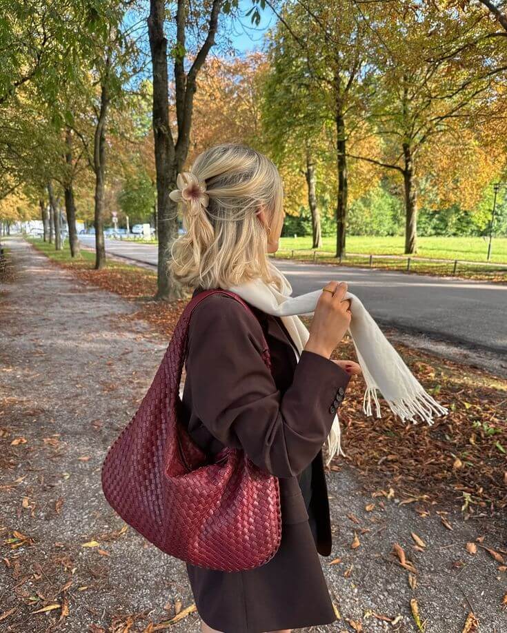 Person holding a red woven bag and white scarf in an autumn park setting

